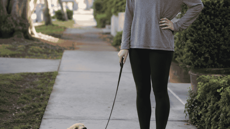 A first-time dog owner standing patiently on a sidewalk while her puppy sits stubbornly and refuses to walk on the leash — one of the most common leash training challenges new owners face when learning how to leash train a puppy